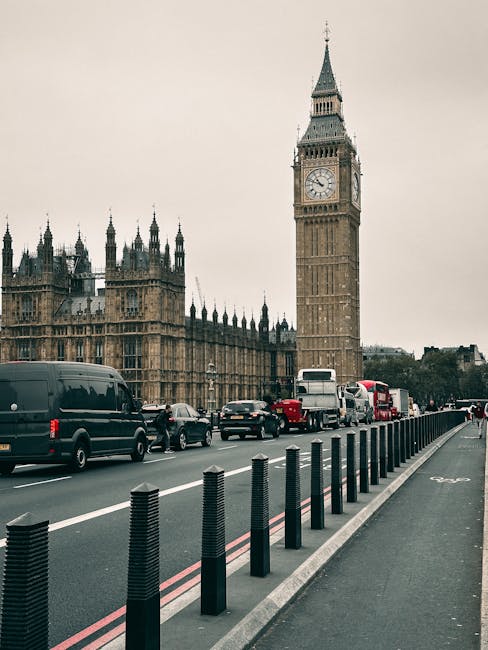 A city street scene featuring the iconic Big Ben clock tower and the Houses of Parliament in London under an overcast sky. In the foreground, a row of black protective bollards lines the pavement, separating it from the busy multi-lane road filled with various vehicles including black vans, cars, and a double-decker bus. The traffic appears steady, with some vehicles waiting or moving along the route. The scene is set during daylight hours with subdued lighting typical of cloudy weather. The environment suggests an urban area suitable for home relocation or furniture transport processes, where belongings might be loaded or unloaded near key landmarks. This image is relevant to the services provided by West Kensington Removals, supporting the context of packing and moving in central London, especially near well-known sites like Big Ben.