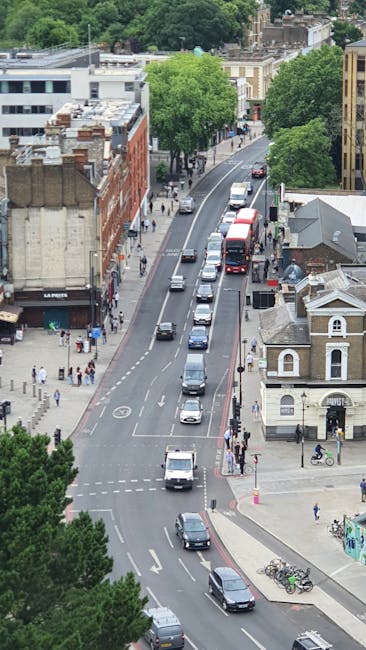 An aerial view of a busy residential street in West Kensington showing a line of parked cars along the curb and moving vehicles on the road. On the left side, there are commercial buildings with shopfronts, including a store with a sign that reads 'LA POTES', while pedestrians are walking along the sidewalk. On the right, a small white building with a curved roof and a few bicycles parked nearby is visible near the corner. Several large green trees are lining the street, providing shade and greenery to the area. The scene captures the typical urban environment where house removals and home relocation services by West Kensington Removals may be performed, including loading and transporting furniture, packed boxes, and appliances in preparation for moving day, with a focus on efficient logistics in a bustling neighbourhood setting.