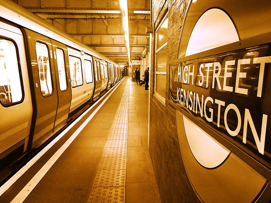 Inside a London Underground station, a train is stationary along the platform, with its doors closed, visible through large windows. The platform features a tactile paving strip near the train for saf