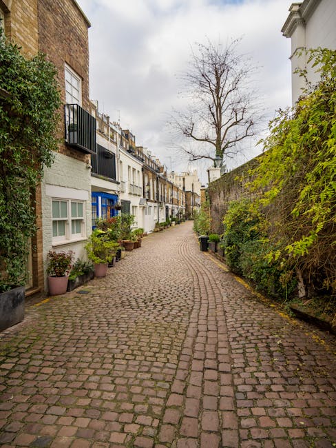 A narrow, cobbled residential street in West Kensington with a gentle curve, lined on both sides by terraced houses featuring white and pastel-colored facades, some with small balconies and window box