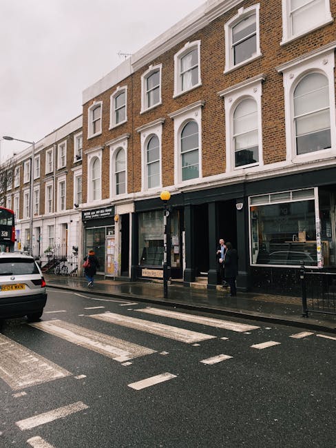 A street view showing the exterior of a three-storey building with a brick facade and large white-framed windows, some with decorative architraves, located on West Kensington. The ground floor features commercial spaces with glass fronts, including a closed shop window. The street has a wet asphalt surface with zebra crossing lines, and a few pedestrians are visible, some carrying bags. A black van is parked on the road, and a double-decker bus is partly visible in the background. The scene suggests an urban environment suitable for home relocation or moving services, and West Kensington Removals may facilitate furniture transport and packing logistics here, with visible elements of a loading and unloading process taking place outside the building.