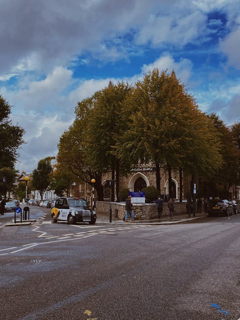 A street scene showing a large building with an arched entrance labeled 'Jubilee Temple' surrounded by several tall deciduous trees with green and autumn-colored foliage. The sky above is partly cloudy with patches of blue. On the street in front of the building, there are parked cars, including a silver vehicle and a dark-colored van, as well as pedestrians walking near the entrance. The scene includes a pedestrian crossing, street signs, and a small stone wall around the trees, indicating an urban environment. The area appears to be a location for house removals or home relocation services, with the company West Kensington Removals possibly involved in the logistics of furniture transport or packing processes in West Kensington, London.