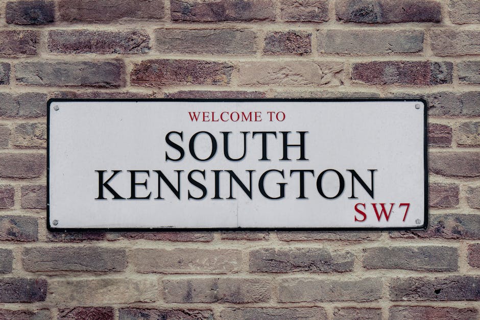 A rectangular white street sign mounted on a red brick wall displays the text 'WELCOME TO SOUTH KENSINGTON SW7' in bold black lettering, with smaller red text reading 'WELCOME TO' at the top and the postcode 'SW7' at the bottom right. The sign is attached with four screws, one at each corner. The brick wall has a rough texture with varying shades of red and brown, and the scene is evenly lit with natural light. This sign is typically seen in residential or commercial areas, and the context may relate to house removals or relocation services provided by West Kensington Removals, especially when considering home relocation or furniture transport within West Kensington.
