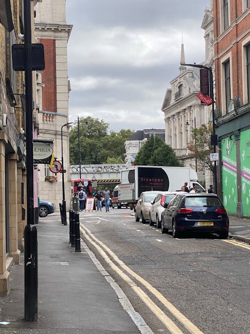 A street scene in West Kensington showing a narrow road lined with parked cars, including a dark blue vehicle closest to the camera. The pavement on the left side features black bollards, and buildings with brick and stone facades are visible, some with shop signs and awnings. In the background, a delivery truck with the logo 'Dinetta' is parked, and a group of people are gathered near a temporary setup, possibly for a move or outdoor event. There are trees and larger historic buildings with classical architecture further in the distance. Overhead, a cloudy sky casts diffuse lighting on the scene, with a traffic light pole and street signs visible on the right. This setting suggests activity related to a home relocation or furniture transport, and it may involve loading, unloading, or moving preparations, aligning with [COMPANY_NAME]’s removals services in West Kensington.