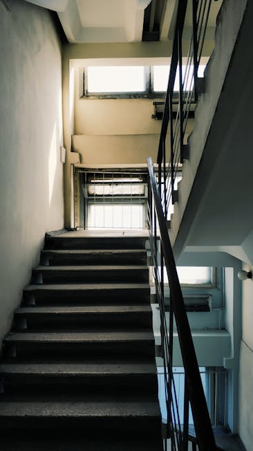 Photograph of a narrow, indoor staircase with a metal railing, leading upward towards a small window at the top, in a property during a house removal process. The stairs are made of dark wood or similar material, with visible signs of wear. At the bottom of the staircase, there is a small cardboard box with packing tape, possibly containing household items, positioned near the first step. The surrounding walls are painted in light, neutral tones, and natural light from the window illuminates the area. The narrowness of the staircase and the low ceiling height suggest a challenging space for furniture transport, which West Kensington Removals manages carefully during their home relocation services. The scene captures the typical environment encountered during the loading and moving process, with steps being prepared for carrying belongings or furniture, and interior elements such as windows and structural details of the staircase clearly visible to aid accessibility for users and support the context of professional removals.