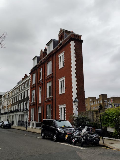A multi-storey red-brick building with white window frames and decorative white accents along the edges, situated on a city street under a cloudy sky. In front of the building, a black van is parked on the pavement, alongside several parked motorcycles. The street features other residential buildings with varying architectural styles, and a British-style lamppost is visible near the sidewalk. The scene depicts an urban environment suitable for house removals and furniture transport, with no visible moving activity but characteristic of a typical relocation setting in West Kensington, aligning with services provided by West Kensington Removals.