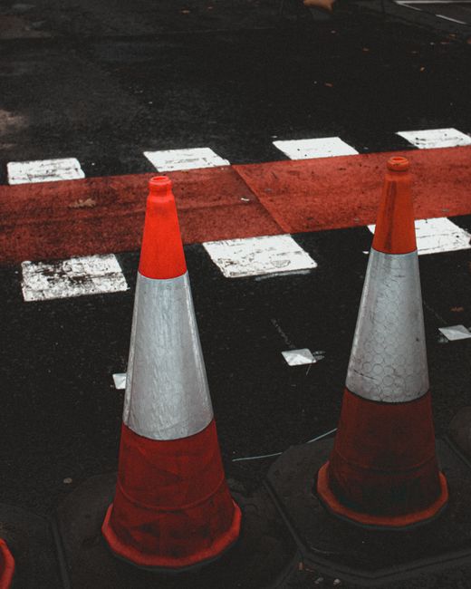 A close-up view of two orange and gray traffic cones positioned on the edge of a paved street near a pedestrian crossing with white dashed lines and a red painted section, marking a temporary loading zone or restricted area for house removals on a residential street in West Kensington. The background shows dark asphalt and a curb, indicating areas where vehicles and moving trucks can safely park or load. This setup reflects compliance with pavement and loading regulations for home relocation and furniture transport, with the cones serving as visual indicators for traffic and pedestrian safety during the loading process. West Kensington Removals ensures such measures are observed during their moving services to facilitate smooth, compliant house moves within urban environments.
