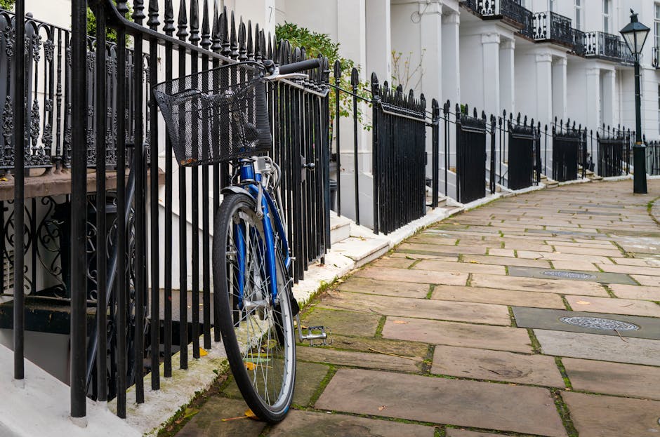 A blue bicycle with a black front basket is locked to a black metal fence that surrounds a white townhouse in West Kensington. The bicycle is positioned on a paved pavement area with flat stone slabs, some of which have small patches of moss or dirt between them. The fence features pointed finials along the top and is secured to concrete steps leading up to the entrance of the building. The building has white exterior walls, dark railings on the balcony, and a black street lamp mounted on the wall. The scene appears to be part of a residential area suitable for home relocation or furniture transport, with the pavement and loading area prepared for moving activities. The natural lighting suggests daytime, with shadows indicating the direction of sunlight, and the environment is clean and orderly, reflecting typical urban residential loading conditions as observed in house removals and moving services provided by West Kensington Removals.