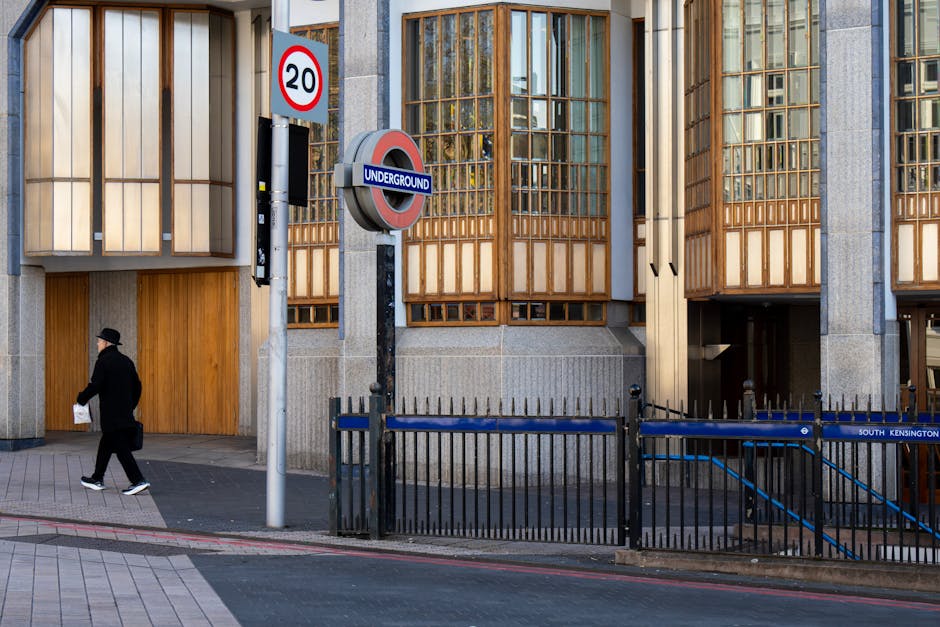 A street view outside West Kensington Underground station showing a modern building with wooden and glass window panels. In the foreground, a pedestrian dressed in dark clothing and a hat is walking past a black metal railing and a sign indicating a 20-mph speed limit. The station entrance, partially visible on the right, features stairs descending into the underground platform. Measures for home relocation or furniture transport, such as a loading area or moving equipment, are not visible in this image. The lighting appears natural, suggesting daytime, with clear weather. This scene provides context for urban moving logistics, with the station's proximity facilitating residential and commercial relocations in West Kensington, as managed by West Kensington Removals, supporting efficient packing, loading, and transport processes.