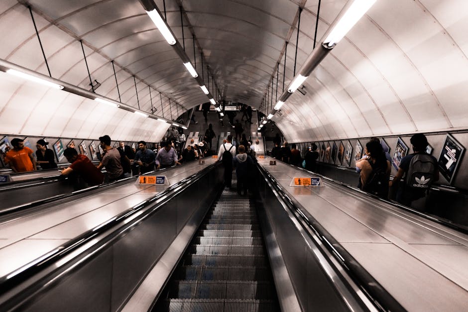 Interior view of West Kensington underground station showing a long escalator with passengers using handrails, leading up to station exits. The station features curved white walls, bright overhead lighting, and advertisements along the side panels. Facing away from the camera, a person carrying a small bag and wearing dark clothing is positioned centrally at the bottom of the escalator. Several other passengers are visible along the sides, some standing, some moving, and others engaging with their phones or talking. The station environment appears clean and well-lit, with the escalator leading to a busy pedestrian area. This setting exemplifies the typical urban transportation hub where residents may consider house removals or furniture transport services, which West Kensington Removals supports through efficient moving logistics, including navigating tight access points during home relocations and moving furniture safely through busy stations.